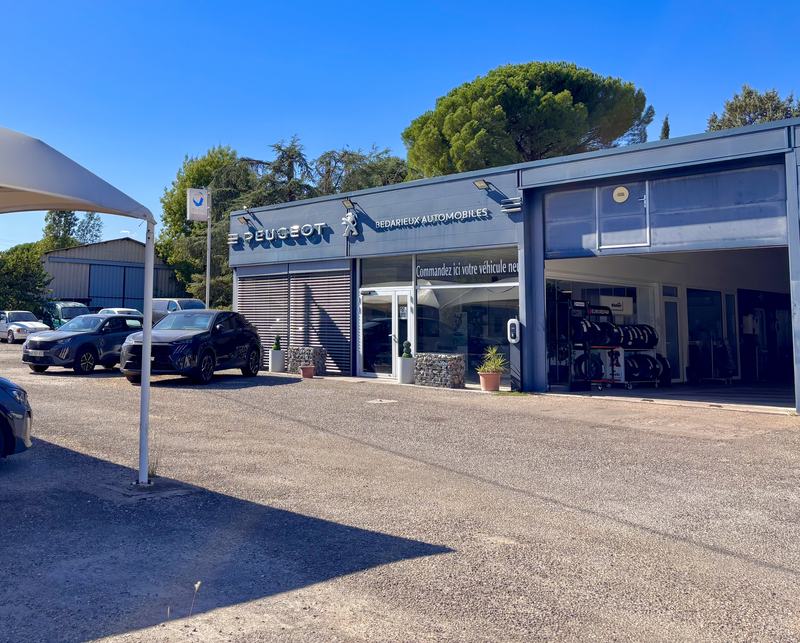 Bâtiment de Bédarieux Automobiles avec l’enseigne Peugeot, parc de véhicules et barnum, sous un ciel bleu